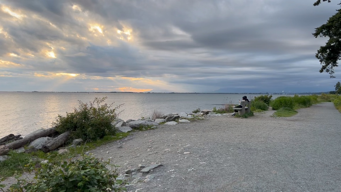 Winding Down by the Beach A coastal view on a cloudy day with golden sunlight breaking through the clouds over the ocean. A gravel walking path runs along the rocky shoreline, where someone is sitting on a bench looking out at the water.