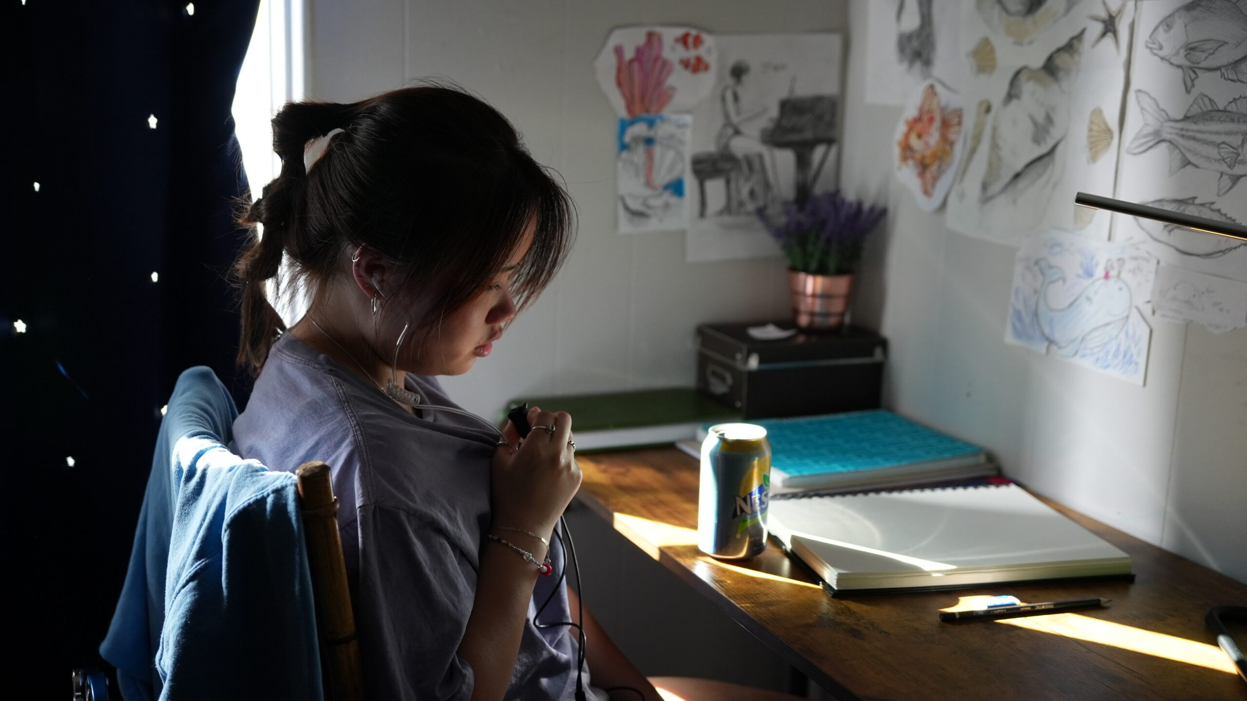 Steffie sits at a sunlit wooden desk, wearing a production headset and holding a clip-on microphone. The wall behind her is decorated with hand-drawn sketches of marine life, including fish and shells, creating a creative and focused atmosphere.