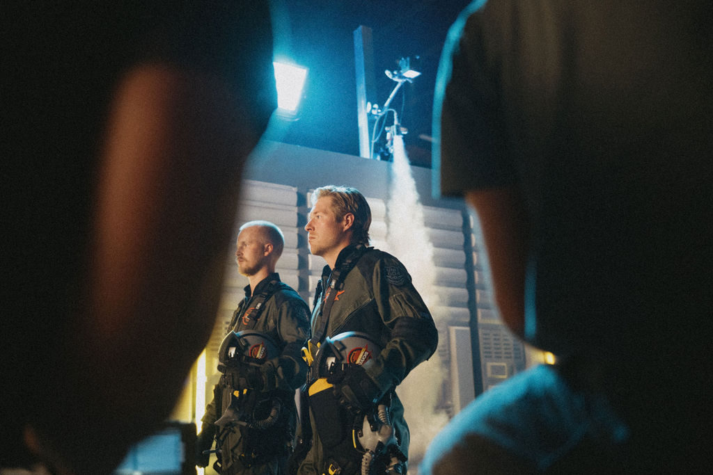 Behind the scenes shot of Elias Pettersson and Brock Boeser in pilot flight gear on a smoke-filled film set.